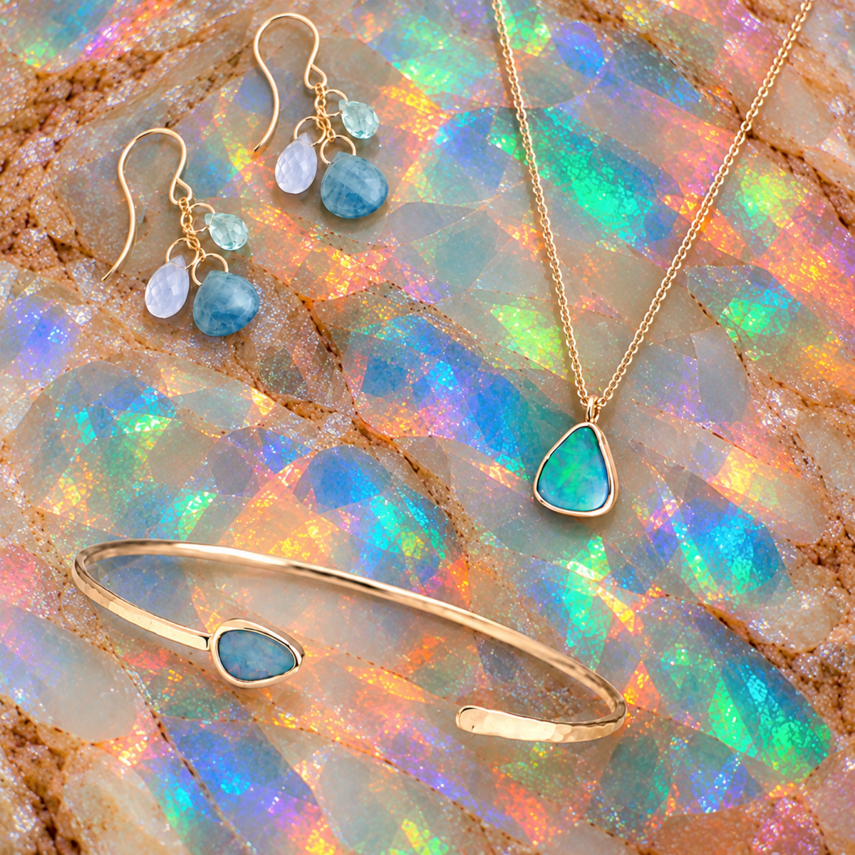 A gold jewelry set featuring a necklace, matching drop earrings, and a cuff bracelet, each adorned with a small light blue stone and a larger teardrop-shaped blue stone, displayed on a colorful opal rock background.